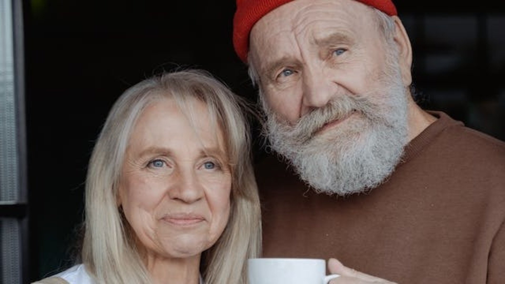 Mann med skjegg og rød lue står sammen med kvinne og han har en kaffekopp i hånden